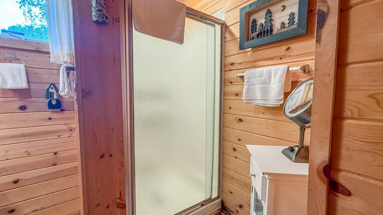 Step into your private bathroom featuring warm wood paneling, a modern shower, and fresh towels ready for your arrival. Natural light and rustic cabin charm create a cozy retreat.