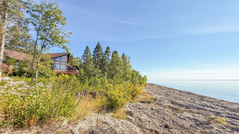 The property sits along pristine lakeside shoreline, surrounded by tall pines and natural rock formations—a peaceful retreat framed by Northern wilderness.
