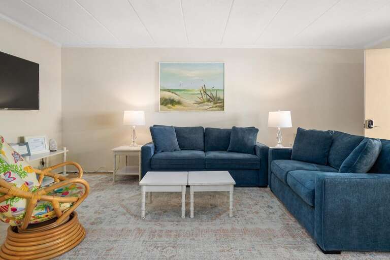 Vacation Rental Living Area With Blue Sofas, White Tables, And Seaside Painting