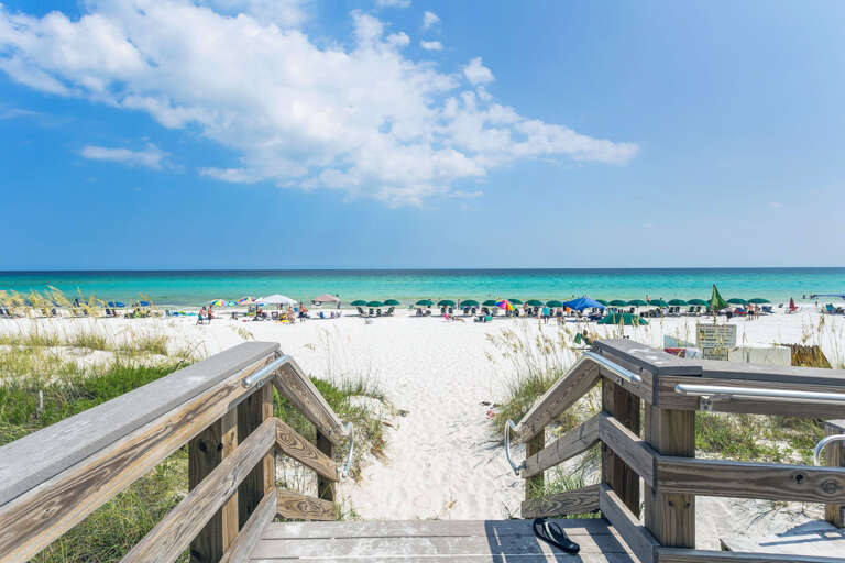 Wooden Boardwalk Leading To Sandy Beach With Umbrellas And Sea In Background
