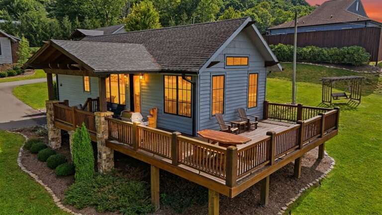 Banner Sky sits on a gentle green lawn with an elevated wraparound deck, Adirondack chairs, and Blue Ridge hills rolling behind.