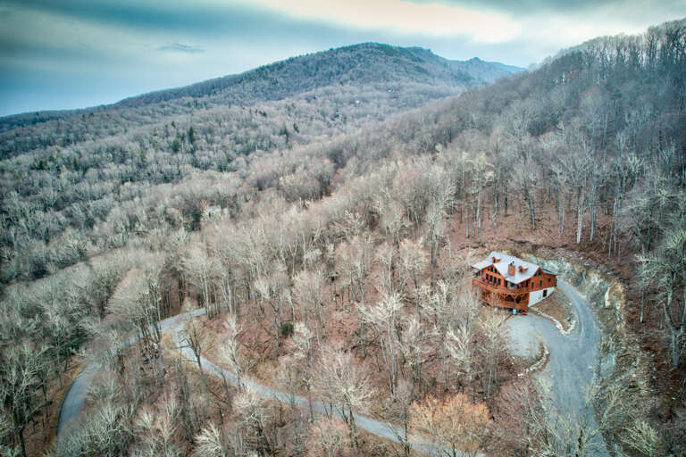 Aerial View Of A Secluded Cabin Surrounded By Frosty, Bare Trees And A Winding Road, Set Against A Hilly Backdrop