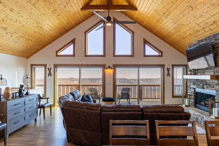Spacious Living Space With Vaulted Wooden Ceilings, Large Windows, And A Stone Fireplace, Overlooking A Scenic Vista