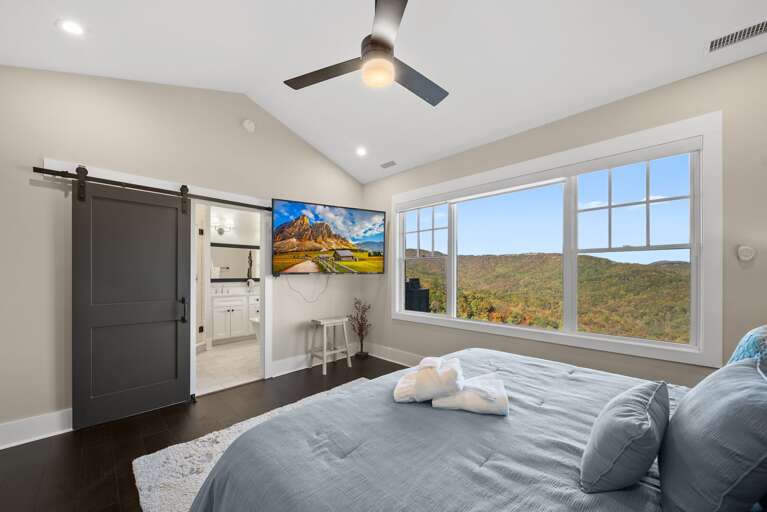 Vacation Rental Room With Rolling Hill View Through Large Windows, Mounted TV, And Ceiling Fan