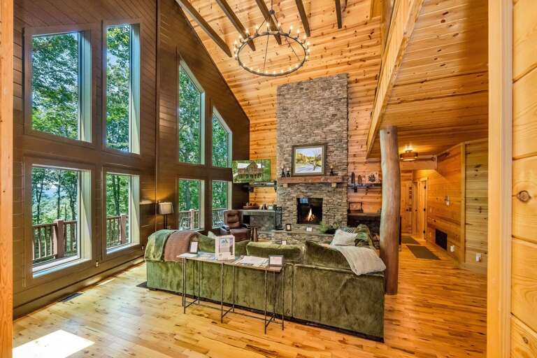 Wood-lined Interior With Towering Windows And Rustic Stone Fireplace Amidst Lush Greenery