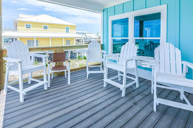 Seaside Scene With White Chairs On Wooden Deck, Bright Blue Beach House Behind Reflecting In Glass Door