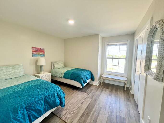 Bright Bedroom With Twin Blue Beds, White Walls, Wooden Floor, And Sunlight Streaming Through A Side Window